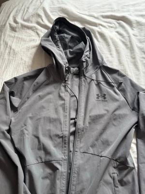Grå vindjacka från Under Armour - Snygg grå vindjacka från Under Armour med huva och dragkedja framtill. Jackan har sportig design, två sidofickor och diskret logga på bröstet. Perfekt för blåsiga dagar och enkel att matcha med andra plagg.