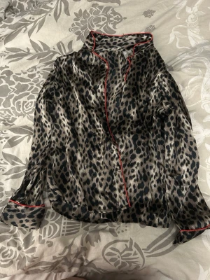 Leopardmönstrad satinpyjamaströja - Snygg långärmad pyjamaströja i glansigt satinmaterial med leopardmönster och röda kontrastkanter. Perfekt för en chill kväll hemma. Skjortmodell med krage och knäppning framtill.