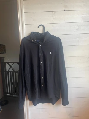 Svart skjorta från Ralph Lauren - Klassisk svart skjorta från Ralph Lauren med vit broderad logga på bröstet. Skjortan har knappar framtill, krage och långa ärmar. Perfekt till en stilren och enkel look.