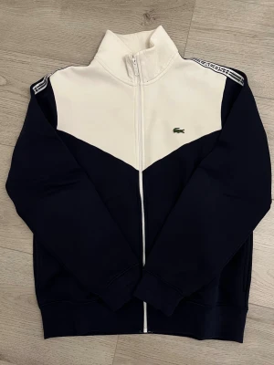 Lacoste ziptröja i marinblått och vitt - Snygg ziptröja från Lacoste i marinblått och vitt med hög krage och dragkedja framtill. Tröjan har ikoniska Lacoste-loggan på bröstet och mönstrade band längs axlarna. Perfekt för en stilren look.