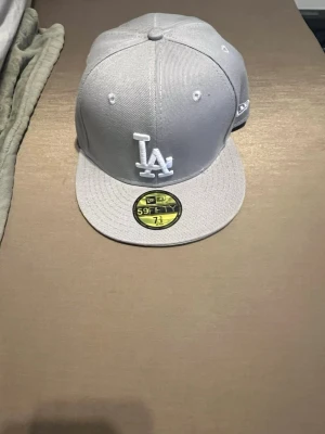 Grå LA New Era 59Fifty keps - Snygg grå New Era 59Fifty keps med LA-logga framtill i vitt broderi. Klassisk flat peak och MLB-logga baktill. Tillverkad i slitstarkt tyg, perfekt för dig som gillar streetwear och sportig stil.