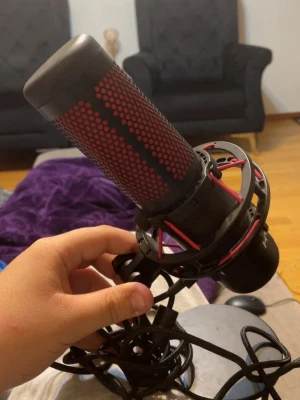 HyperX röd och svart mikrofon - Säljer en HyperX mikrofon i svart och rött med metallgaller och stötdämpande upphängning. Perfekt för streaming, gaming eller poddande. USB-anslutning och stabil rund fot. Snygg design med tydlig HyperX-logga framtill.