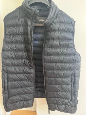 Svart dunväst från Dressmann - Snygg svart dunväst från Dressmann med quiltad design och hög krage. Västen har dragkedja framtill och två sidofickor. Perfekt att slänga över en hoodie eller tröja för extra värme. Tillverkad i lätt och smidigt material.