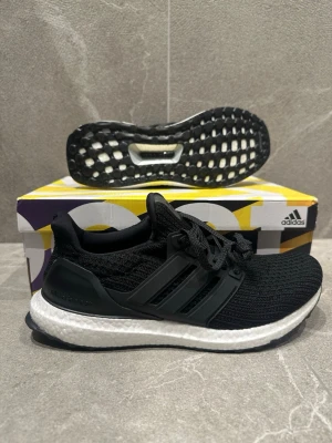 Adidas Ultraboost svarta sneakers - Svarta Adidas Ultraboost sneakers med stickad ovandel i mesh och klassiska tre ränder på sidan. Skon har en vit Boost-mellansula för extra komfort och svart yttersula med grepp. Perfekt för dig som gillar stilrena och sportiga sneakers.