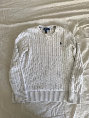 Vit kabelstickad tröja Polo Ralph Lauren - Kabelstickad vit tröja från Polo Ralph Lauren med rund halsringning och ribbade muddar. Klassisk marinblå logga broderad på bröstet. Perfekt till jeans eller kjol för en clean och tidlös look. Pris kan diskuteras 