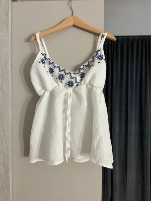Vit linnetopp med blå broderi - Söt vit linnetopp med smala axelband och blå broderade blommor längs ringningen. Toppen har en v-ringad form och knytdetalj framtill. Perfekt för varma dagar och ger en fräsch sommarkänsla.