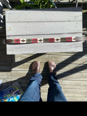 Pampeano bälte - Snyggt bälte i brunt skinn med färgglada blockmönster i rött, grönt, svart och vitt. Bältet har ett klassiskt spänne i metall och ett unikt geometriskt mönster längs hela bandet. Perfekt för att ge din outfit en cool touch.