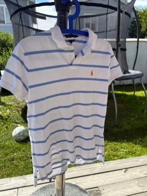 Vit och blårandig pikétröja Polo Ralph Lauren - Klassisk vit pikétröja från Polo Ralph Lauren med ljusblå ränder och den ikoniska loggan broderad på bröstet. Tröjan har korta ärmar, krage och knappar vid halsen. Perfekt för en avslappnad och stilren look.