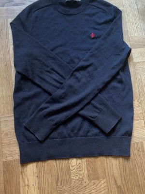 Mörkblå stickad tröja från Morris - Mörkblå stickad tröja från Morris i 100% merinoull. Tröjan har rund halsringning, långa ärmar och en liten röd broderad logga på bröstet. Ribbstickade muddar vid ärmslut och nederkant ger en snygg passform.