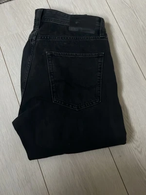 Svarta jeans från Jack & Jones - Säljer ett par svarta jeans från Jack & Jones med klassisk femficksmodell och rak passform. Jeansen har en diskret logopatch bak i midjan och är tillverkade i ett mjukt denimtyg som är sköna att bära. Perfekta till en avslappnad stil.