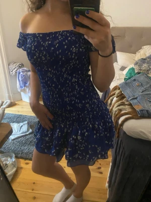 Blå offshoulder blommig kortklänning - Söt blå kortklänning med vita små blommor och offshoulder-design. Klänningen har volangkant nedtill och smockad överdel som ger en figurnära passform. Perfekt för varma dagar och sommarens alla tillfällen.