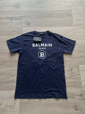Marinblå Balmain Paris t-shirt - Snygg marinblå t-shirt från Balmain Paris med vit logotyp och tryck på bröstet. Klassisk passform och rund halsringning, perfekt för dig som vill ha en stilren och trendig look. Materialet är mjuk bomull som känns skönt mot huden.