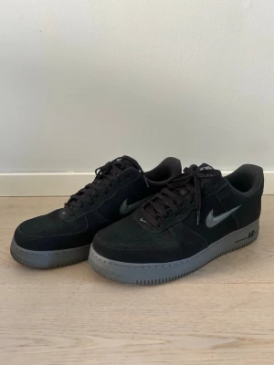 Nike Air Force 1 svart/silver sneakers - Snygga Nike Air Force 1 sneakers i svart mocka med silverfärgad swoosh och grå sula. Klassisk låg modell med snörning och tydlig Nike-logga på sidan. Perfekta för dig som gillar stilrena och tidlösa sneakers.
