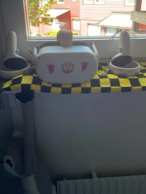 Meta Quest 2 VR-headset med kontroller - Säljer ett vitt Meta Quest 2 VR-headset med två matchande handkontroller. Headsetet är dekorerat med Manchester United-klistermärken på framsidan. Tillverkad i plast med en modern och stilren design, perfekt för gaming och VR-upplevelser.