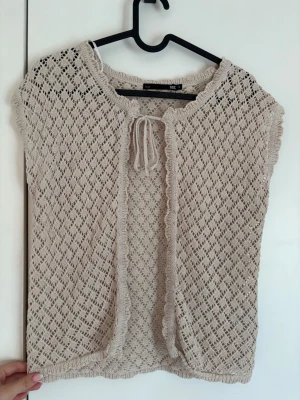 Beige stickad väst🫶🏻 - Söt beige stickad väst från Lager 157 med korta ärmar och knytband framtill. Västen har ett luftigt hålmönster och volangkant längs öppningen. Perfekt att slänga över ett linne för en trendig lager-på-lager-look. Jätte gullig!!🤍
