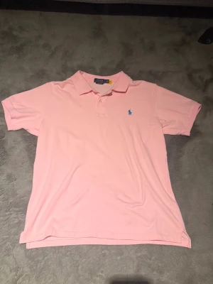 Rosa pikétröja från Ralph Lauren - Säljer en klassisk rosa pikétröja från Ralph Lauren med blå logga broderad på bröstet. Tröjan har korta ärmar, krage och knappar framtill. Perfekt för dig som gillar stilrena och tidlösa plagg.