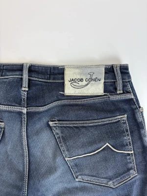 Mörkblå slim fit jeans Jacob Cohen - Snygga mörkblå slim fit jeans från Jacob Cohen med klassisk femficksmodell och kontrastsömmar. Jeansen har en tydlig logotyp-patch bak och subtila slitningar för en trendig look. Perfekta för dig som gillar stilrena och moderna jeans.