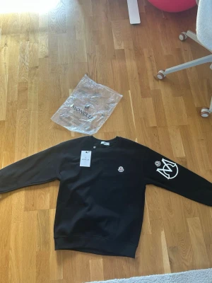 Svart sweatshirt från Moncler - Snygg svart sweatshirt från Moncler med rund hals och ribbade muddar. Tröjan har Moncler-logga på bröstet och ett stort vitt tryck på ena ärmen. Tillverkad i mjukt bomullsmaterial som känns skönt mot huden. Perfekt för en trendig och avslappnad stil. Pris ej hugget i sten.