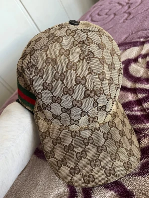 Gucci keps - Snygg beige keps från Gucci med klassiskt GG-monogram över hela kepsen. På sidan finns de ikoniska gröna och röda ränderna. Kepsen har böjd skärm och justerbar passform. Perfekt accessoar för att lyfta din streetwear-look.
