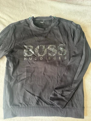 Svart sweatshirt från Hugo Boss - Svart långärmad sweatshirt från Hugo Boss med stort BOSS-tryck och logga framtill. Tröjan har rund halsringning och ribbade muddar vid ärmslut och nederkant. Perfekt för en stilren och avslappnad look.