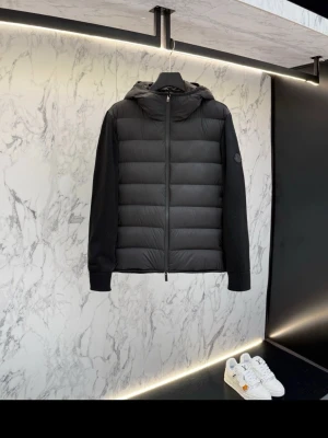 Svart pufferjacka från Moncler - Snygg svart pufferjacka från Moncler med quiltad front och rygg, samt stickade ärmar och huva. Jackan har dragkedja framtill och Moncler-logga på ärmen. Perfekt för kalla dagar när du vill hålla stilen.