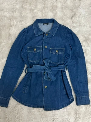 Jeansjacka med puffärm och bälte – Kappahl – Stl S - Säljer en jättefin jeansjacka från Kappahl i en snygg mörkare blå denim. Jackan har en feminin design med dekorativa puffärmar vid axlarna och ett matchande knytband i midjan som ger en fantastisk siluett. Super fin och fungerar utmärkt som vårjacka!