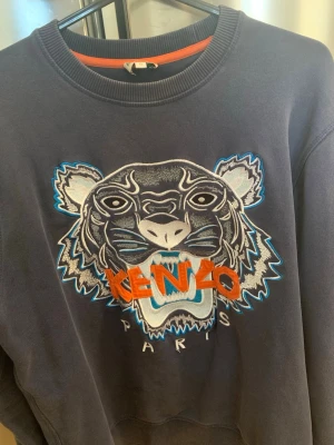 Kenzo sweatshirt med tigertryck - Mörkblå sweatshirt från Kenzo med ikoniskt broderat tigerhuvud och färgglad logga på bröstet. Rund halsringning, ribbade muddar och avslappnad passform. Perfekt statement-plagg för dig som gillar streetwear och snygga detaljer.