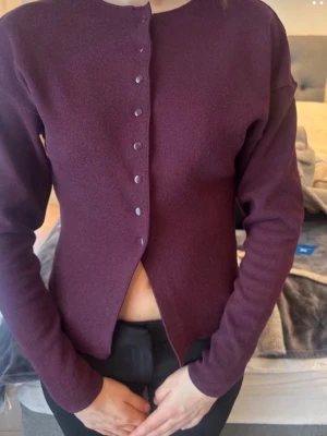 Mörklila kofta med knappar - Snygg mörklila kofta med rund halsringning och matta knappar framtill. Modellen är figurnära med långa ärmar och lätt stretchigt material. Perfekt att styla till både jeans och kjol för en trendig look.