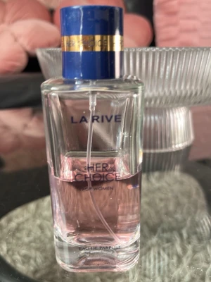 La Rive Her Choice Eau de Parfum - La Rive Her Choice Eau de Parfum för kvinnor i en stilren, rektangulär glasflaska med blått och guldigt lock. Parfymen har en ljusrosa nyans och flaskan är transparent vilket ger en fräsch och modern känsla.