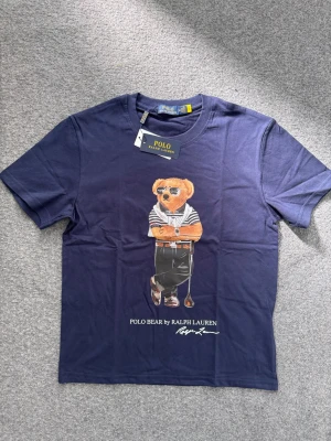 Blå Polo Bear t-shirt från Ralph Lauren - Snygg mörkblå t-shirt från Ralph Lauren med Polo Bear-tryck framtill. Klassisk passform med korta ärmar och rund hals. Tillverkad i mjuk bomull för skön känsla. Perfekt för sommaren och den varma luften! OBS: Polo Ralph Lauren påse ingår i köpet (skickar bild privat), samt kan priset diskuteras! 