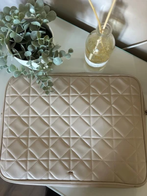 beige datorväska från Åhléns - Stilren datorväska i beige quiltat konstläder från Åhléns. Väskan har dragkedja runtom och ett diskret metallogo framtill. Perfekt för att bära din laptop med en trendig touch och skydda den mot repor. defekter inuti fodralet, då de legat pennor😆men syns inte utanpå fodralet & annars i mycket bra skick