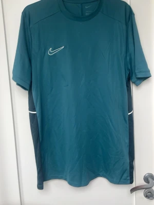 Nike blågrönt träningsset - Säljer ett blågrönt träningsset från Nike med t-shirt och shorts. Båda plaggen har vita Nike-loggor och svarta sidopaneler med vita detaljer. Tillverkade i lätt och ventilerande funktionsmaterial, perfekt för träning eller sport.