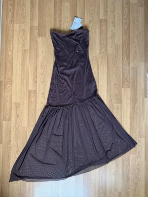 Brun långklänning med paljetter Gina Tricot - Snygg brun långklänning från Gina Tricot med glittriga paljettdetaljer. Klänningen är axelbandslös och har en figurnära passform upptill med ett utsvängt, voluminöst nedre parti. Tillverkad i återvunnen polyester.