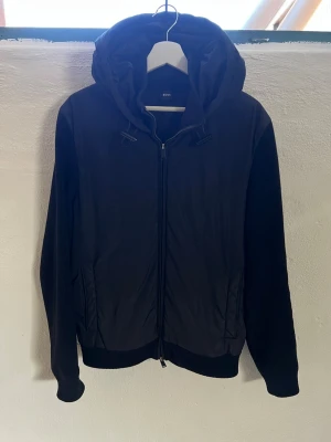 Cardigan Hugo boss X Porsche  - Svart cardiganjacka från Boss med huva och dragkedja framtill. Jackan har ribbstickade ärmar och sidor, samt slät front och ryggparti upptill. Praktiska fickor och sportig look, perfekt för lager-på-lager-stil. Collab med Porsche så väldigt svår och få tag . Speciellt i denna modell 