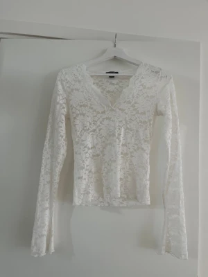 Vit spetsblus med v-ringning - Superfin vit blus i spets med v-ringad hals och långa ärmar. Blusen har ett blommigt spetsmönster och är genomskinlig, perfekt att styla med ett linne under. Snygg och feminin look som passar till många outfits.