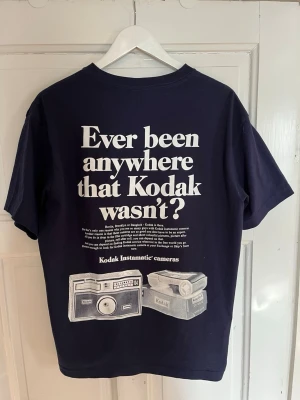 Mörkblå Kodak t-shirt med tryck - Cool mörkblå t-shirt från Kodak med stort retrotryck på ryggen. Trycket har texten 'Ever been anywhere that Kodak wasn’t?' och motiv av gamla kameror. T-shirten har rund hals och korta ärmar, perfekt för dig som gillar vintage och foto.