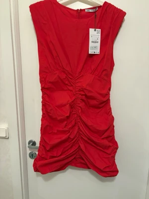 Röd ruched klänning från Zara - Snygg röd kortklänning från Zara med ruched detaljer längs hela framsidan. Klänningen är ärmlös och har en figurnära passform som framhäver siluetten. Perfekt för dig som vill sticka ut med en färgstark look.