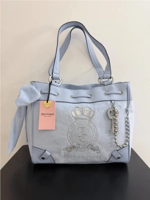  Juicy Couture daydreamer tote  - Superfin ljusblå handväska från Juicy Couture med broderad logga framtill, stora handtag och dekorativ rosett på sidan. Väskan har silverfärgade detaljer, nyckel- och pärlberlock samt är gjord i mjukt tygmaterial. Perfekt för dig som gillar en söt och trendig look.