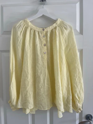 Gul blus  - Ljusgul, luftig blus i oversized modell med rundad halsringning och fyra knappar framtill. Tillverkad i ett tunt, skrynkligt tyg som ger en avslappnad känsla. Långa ärmar med knapp vid ärmslut. Perfekt för en trendig look.