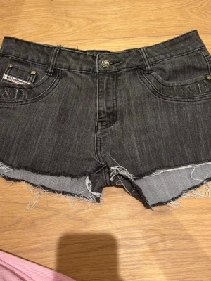 Svarta jeansshorts från D&D - Säljer ett par svarta jeansshorts från D&D med råa, fransiga kanter och klassisk femficksdesign. Shortsen har broderad logga på fickan och detaljer i metall. Perfekta för en avslappnad och cool stil. Lappen på baksidan är trasig därav priset. Men utöver det inga defekter 
