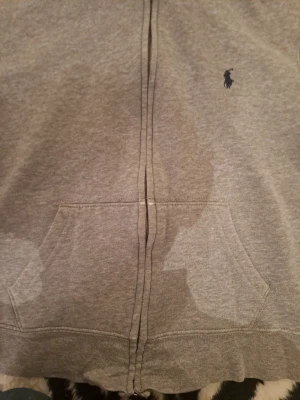 Grå hoodie från Polo Ralph Lauren - Klassisk grå hoodie från Polo Ralph Lauren med dragkedja framtill och kängurufickor. Liten broderad logga på bröstet. Tillverkad i mjukt bomullsmaterial och har ribbade muddar vid ärmslut och nederkant. Perfekt för en avslappnad och stilren look.