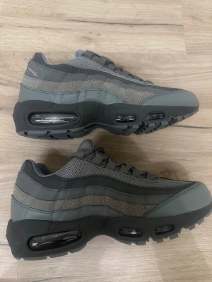Nike Air Max 95 grå sneakers - Snygga Nike Air Max 95 sneakers i olika nyanser av grått med svarta detaljer. Skorna har synliga Air Max-bubblor i sulan, meshpaneler och mockaliknande material på ovandelen. Perfekta för dig som gillar en sportig och stilren look.