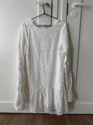 Vit broderad kortklänning med volang - Supersöt vit kortklänning med broderade blommor, volangkant nedtill och långa ärmar. Klänningen har öppen rygg med knytband i nacken och är gjord i ett lätt, luftigt material. Perfekt för sommarens alla tillfällen!