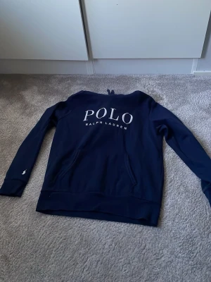 Mörkblå hoodie från Polo Ralph Lauren - Snygg mörkblå hoodie från Polo Ralph Lauren med stor vit logga på bröstet och klassisk broderad polospelare på ärmen. Hoodien har känguruficka och ribbade muddar. Perfekt för en avslappnad och stilren look. Använd enstaka gånger, växt ur väldigt snabbt. 