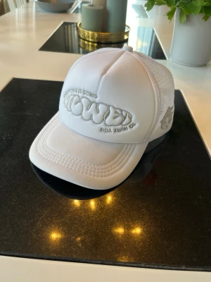 Vit truckerkeps - Säljer en vit truckerkeps med broderad logga framtill och mesh på sidorna för extra ventilation. Kepsen har böjd skärm och justerbar snapback baktill. Perfekt för dig som vill ha en clean och stilren look.