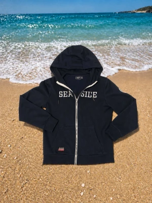 Seaside Mörkblå Tröja - Snygg marinblå hoodie från Seaside med vit dragkedja och tryckt text på bröstet. Hoodien har huva med dragsko, två fickor framtill och ribbade muddar vid ärmar och nederkant. Perfekt för chill dagar vid stranden eller stan.