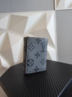 Grå plånbok - Snygg grå plånbok med klassiskt monogrammönster i svart. Plånboken är tillverkad i strukturerat läder och har en kompakt, rektangulär form. Perfekt för att organisera kort och kontanter med stil.