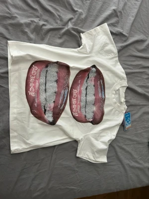 Vit t-shirt med grafiskt tryck - Vit t-shirt med stort grafiskt tryck av två läppar på framsidan. T-shirten har rund halsringning och korta ärmar. Materialet känns som bomull och passformen är normal. Perfekt för dig som vill sticka ut med en unik design.