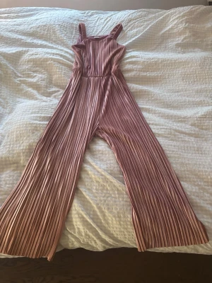 Plisserad rosa byxdress från Lindex - Säljer en gullig plisserad byxdress från Lindex i en mjuk rosa nyans. Byxdressen har breda axelband, resår i midjan och vida ben. Materialet är glansigt och ger en lyxig känsla. Perfekt för er som har mindre barn💕