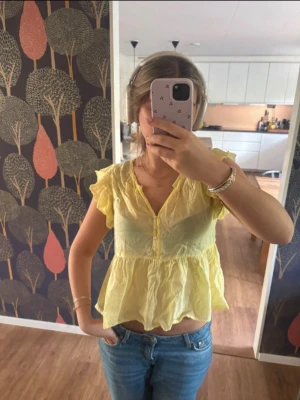 Gul volangblus med v-ringning - Söt gul blus från Mango med volangdetaljer och v-ringning. Aldrig använd, har prislappen kvar. Blusen har korta ärmar, knappar framtill och är tillverkad i ett tunt, luftigt material som passar perfekt till sommaren. Den har en lös och flowy passform.